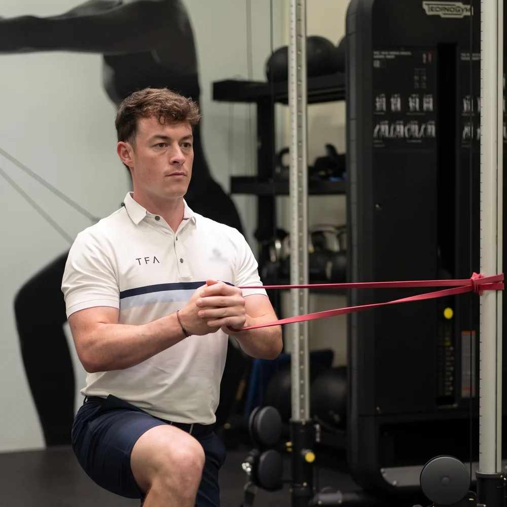Jamie in his TFA polo, holding a band from half kneeling in the academy gym